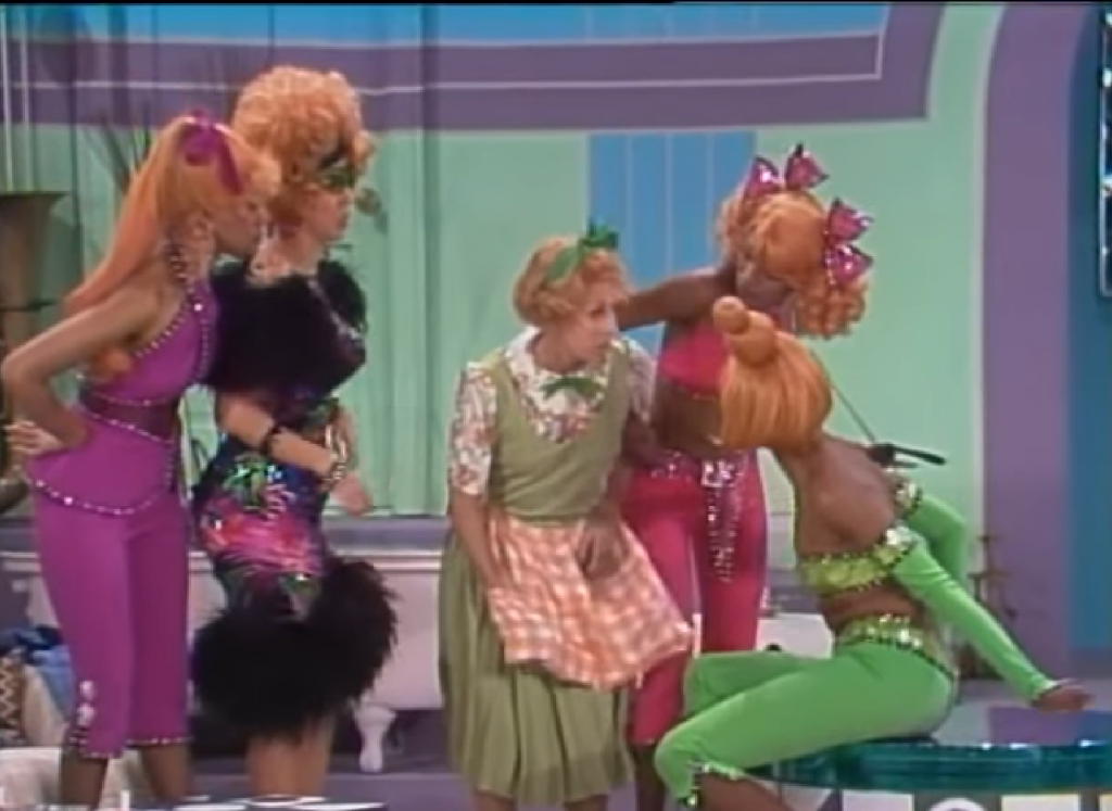 A frumpy Cinderella surrounded by her stepmother, dressed in a fluffy black gown, and her three stepsisters in purple, green, and pink seventies-style cropped pantsuits. 